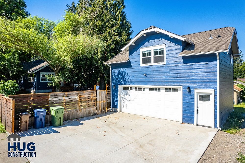 A blue two-story detached garage with Accessory Dwelling Units next to a driveway with recycling bins, surrounded by trees.