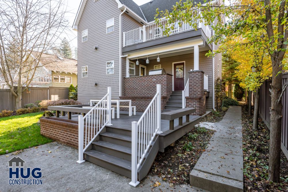 A residential two-story home with a wooden deck and brick stairway leading to a white-railed porch, surrounded by a well-maintained yard with autumn foliage, featuring remodels and additions.