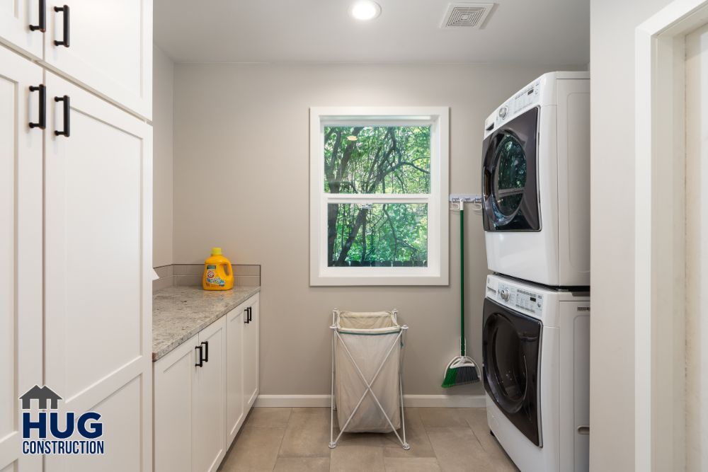 Modern laundry room remodels with stacked washer and dryer, white cabinetry, and a view of greenery through the window.