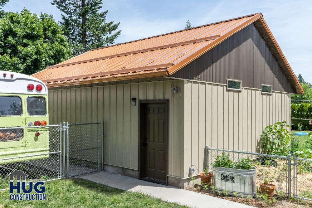 A beige shed with a brown metal roof and a single door, featuring remodels and additions, is situated next to a chain-link fence and greenery, with a partial view of a bus on the