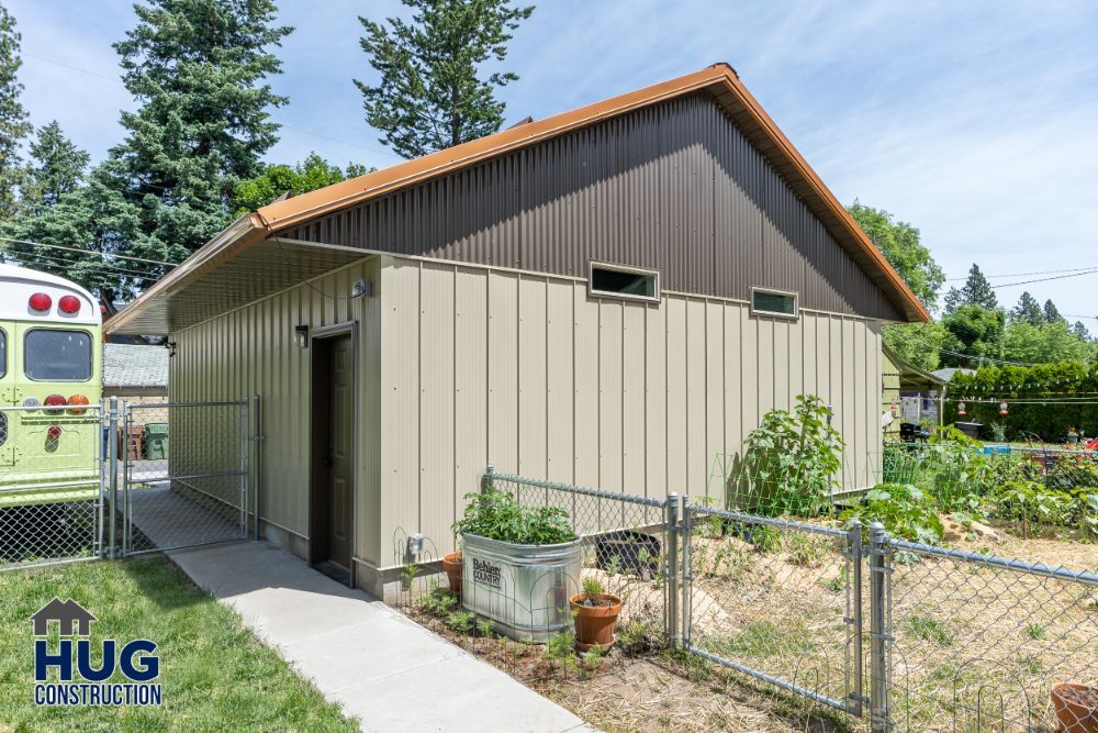 A beige shed with a brown roof, featuring remodels and additions, in a fenced garden area, with a bus visible to the left.