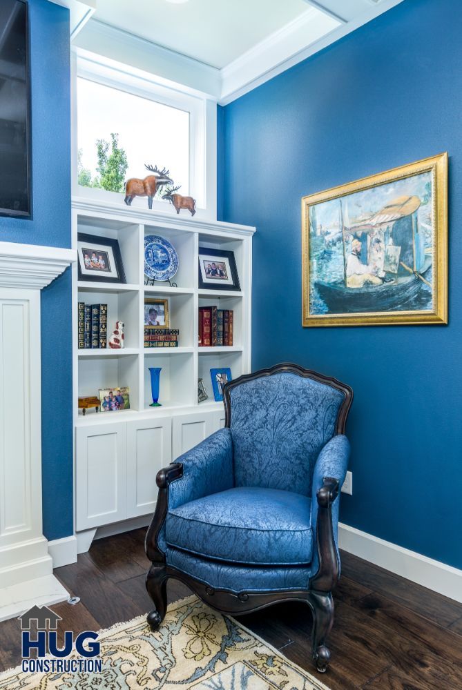 A cozy reading nook, recently remodeled with a blue upholstered armchair, white built-in bookshelves, and dark hardwood flooring, accented by a blue wall and decorative elements.