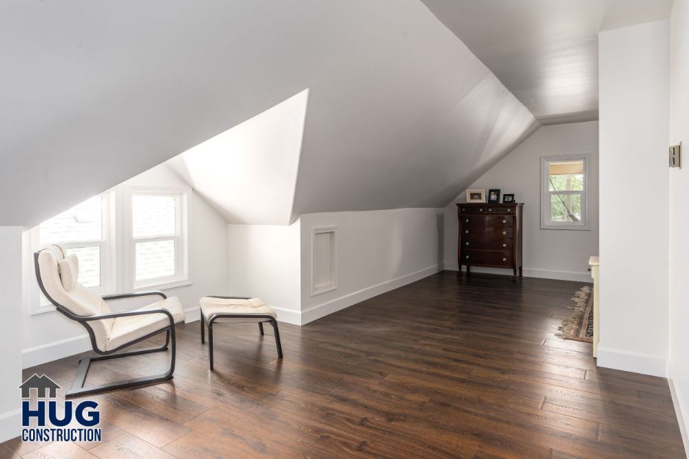 A bright, spacious attic room with hardwood flooring and a slanted ceiling, remodeled and furnished with a chair, an ottoman, and a dark wooden cabinet.