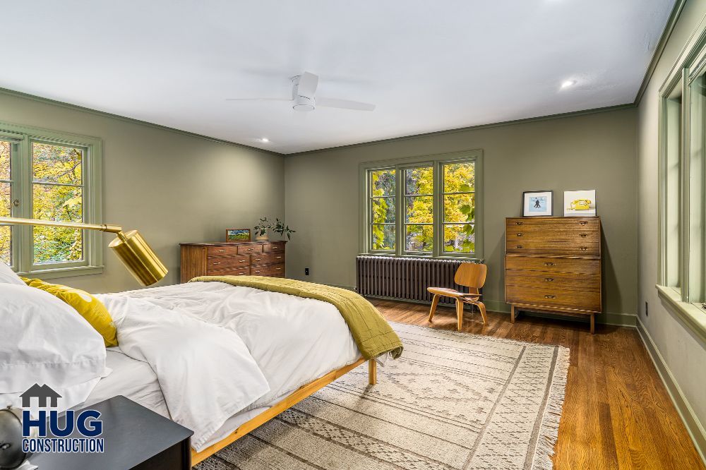 A neatly furnished bedroom with a bed, dresser, and a desk by the window, showcasing ample natural light and green color scheme, enhanced by thoughtful remodels.