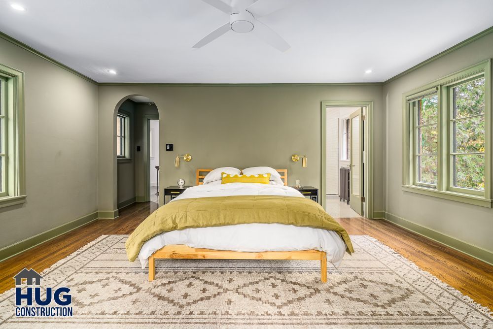 Spacious bedroom remodels with green walls, hardwood floors, and a neutral area rug, featuring a bed with yellow pillows and a ceiling fan above.
