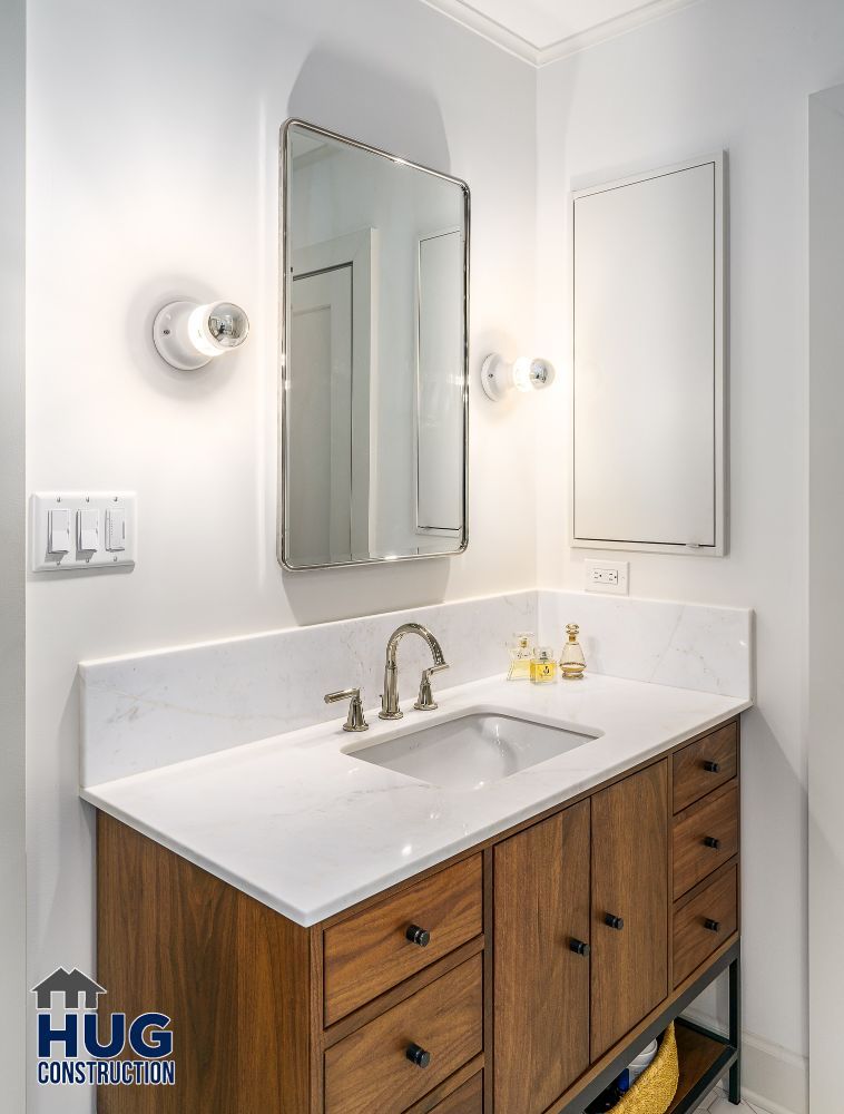 Modern bathroom remodels with a wooden vanity unit, white countertop, wall-mounted mirror, and sconce lighting.