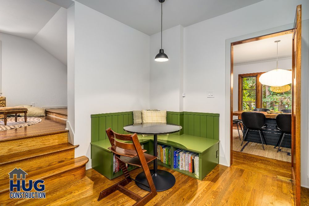 A cozy corner dining nook with built-in green bench seating and a round table, resulting from recent remodels, is adjacent to a living space with hardwood floors.