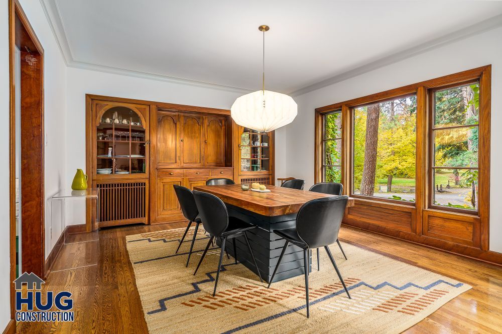 A bright dining room showcasing remodels and additions, featuring a modern table with black chairs, traditional wooden cabinetry, and large windows with a view of greenery outside.