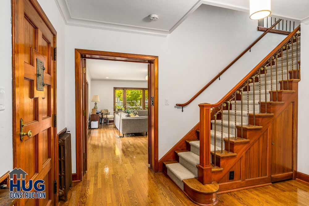 Interior view of a home featuring polished wooden floors, a staircase with wooden banisters, and an open doorway leading to a living room with recent remodels and additions.