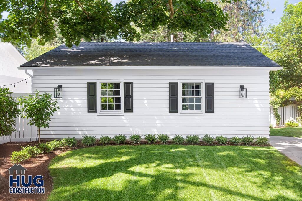 A small, neatly-maintained white house with black shutters under a green tree with a manicured lawn, featuring remodels and additions, and a logo for hug construction at the bottom.