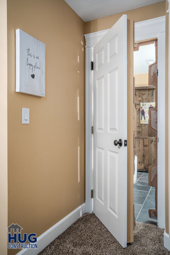 Interior view of a home with remodels and additions, featuring an open white door leading to a bathroom, a closed wooden door in the background, beige walls, and a decorative sign hanging on