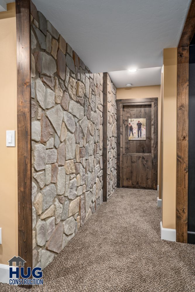 A corridor with stone walls, wooden accents, and carpeted flooring leading to a wooden door with a framed picture hanging above it, showcasing recent remodels and additions.