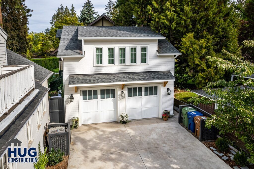 A two-story detached garage with white siding and dual white garage doors, featuring a shingled gable roof, with a landscaped perimeter and a concrete driveway for all projects.