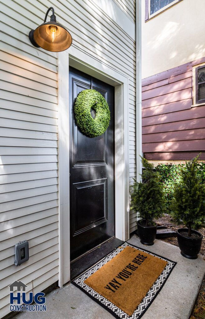 A residential entrance featuring a black front door adorned with a green wreath, flanked by potted plants, with an "All Projects" welcome mat in front of the door, all illuminated by an