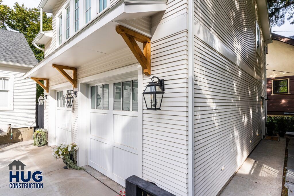 Exterior view of a house showcasing white siding, a garage door, and wooden braces supporting an overhang, with a logo of Hug Construction in the corner. All Projects