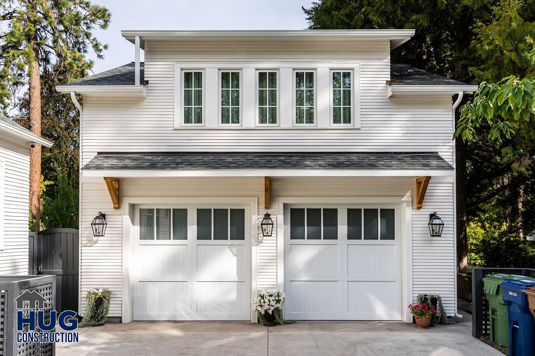 A two-story white house with a double garage door flanked by potted plants and All Projects recycling bins to the side.