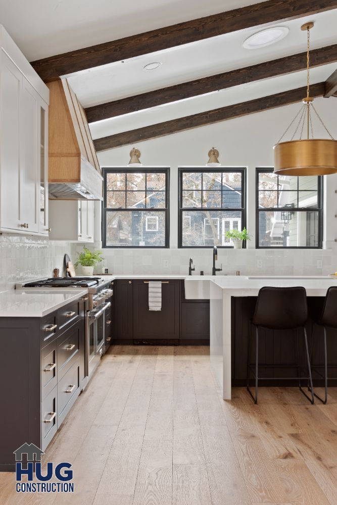 Modern kitchen remodels featuring white cabinetry, dark countertops, exposed wooden beams, and a gold light fixture, with a central island and bar stools.