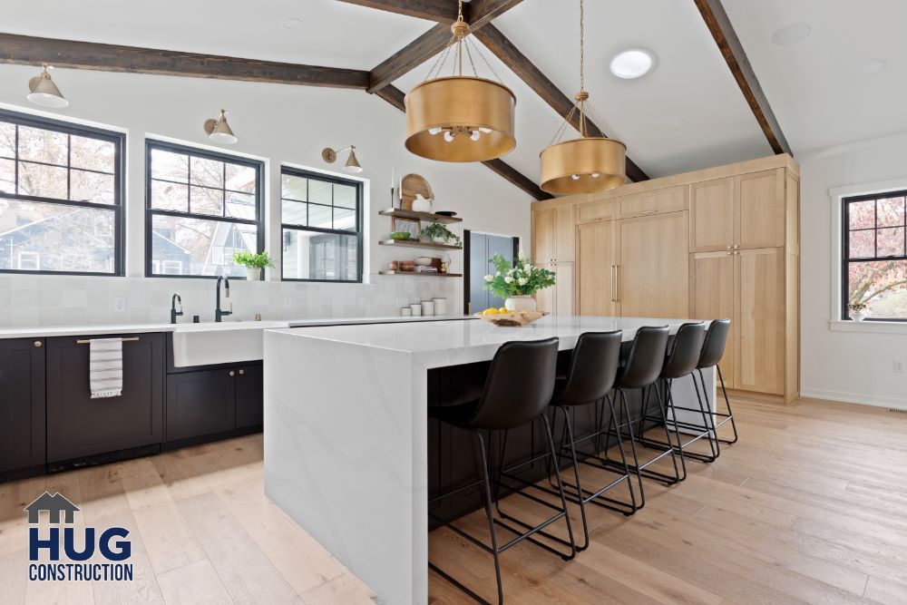 Modern kitchen interior with a large island, remodels, black bar stools, wooden beams, and brass pendant lights.