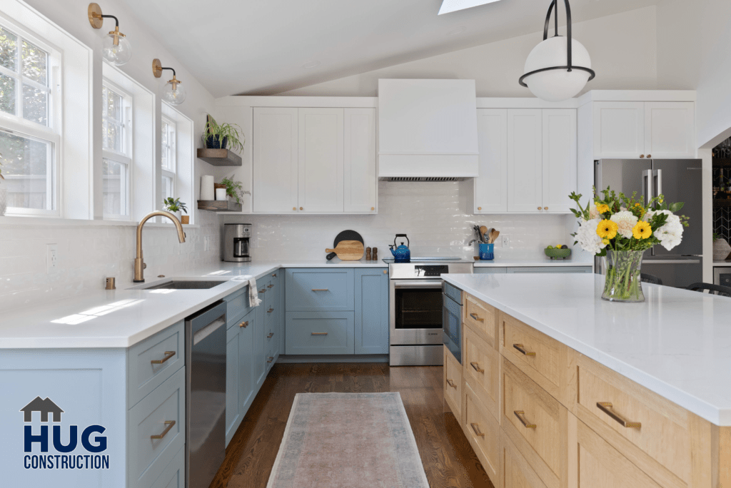 A modern kitchen expansion featuring white upper cabinets, blue lower cabinets, wooden countertops, stainless steel appliances, and a central island under pendant lights. The addition of a bouquet of yellow flowers adds a splash of
