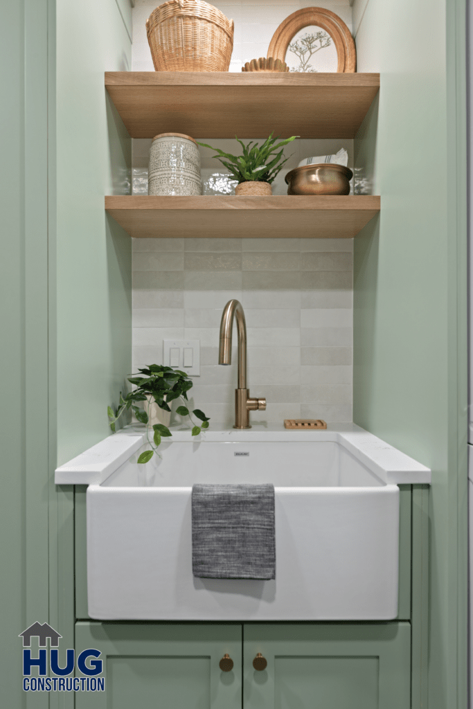 A neatly arranged modern bathroom vanity, part of the Gunning Rd Interior Remodel, with a white rectangular basin, brass faucet, and wooden shelving containing decorative items and plants, against a tiled wall