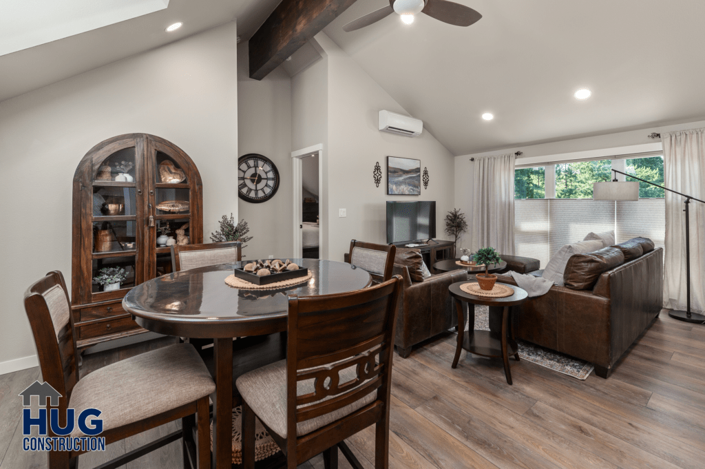 A modern living room featuring a dining area with a round wooden table and chairs, and a lounge area with a brown leather sofa, complemented by a neutral color palette and wooden flooring. The room,