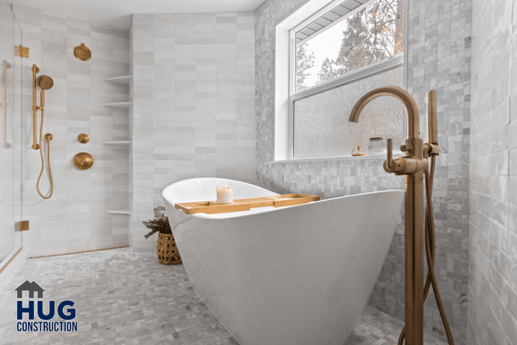 Modern bathroom interior from the Gunning Rd Interior Remodel with a standalone white bathtub, gold fixtures, and neutral tiled walls. A window provides natural light, and a shelf with decorative items adds a touch