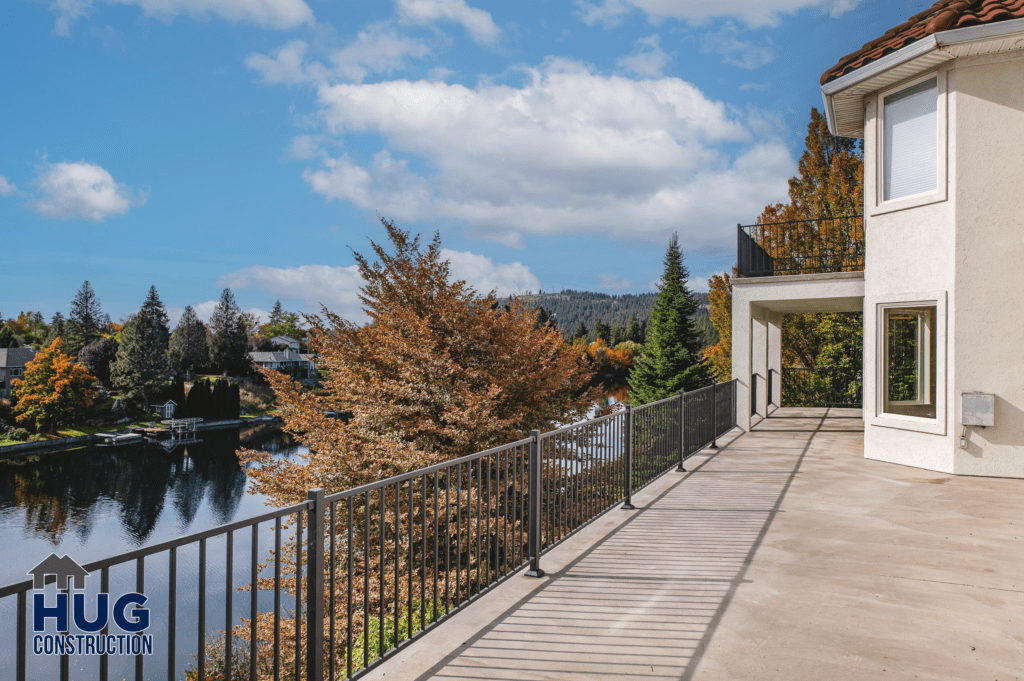 A spacious terrace of a modern home, part of a 2-story deck rebuild with a metal railing, offering a clear view of a serene lake and autumn-colored trees under a partly cloudy sky.