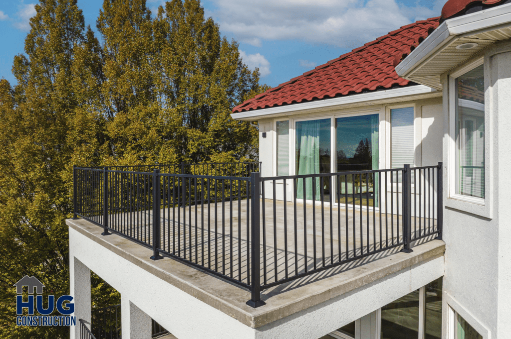 A modern 2-story deck with a black railing, attached to a house with a red tiled roof on a sunny day with trees in the background.