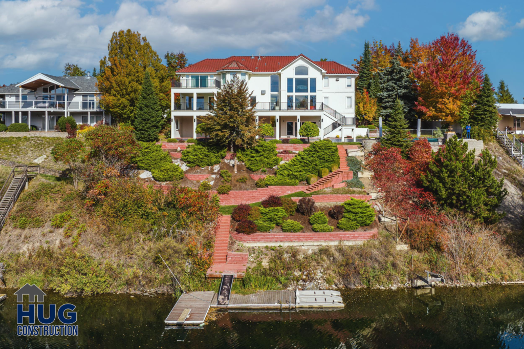 Large white 2-story residential building with blue roof overlooking a water body, featuring terraced landscaping with red-brick pathways and a variety of trees in autumn colors. A small deck extends into the water