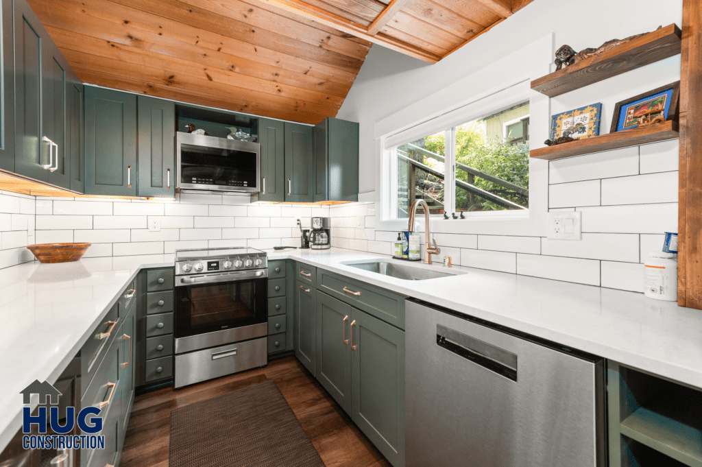 A modern kitchen in the Silver Beach Cabin remodel, with stainless steel appliances, white subway tile backsplash, and dark green cabinetry under a wooden ceiling.