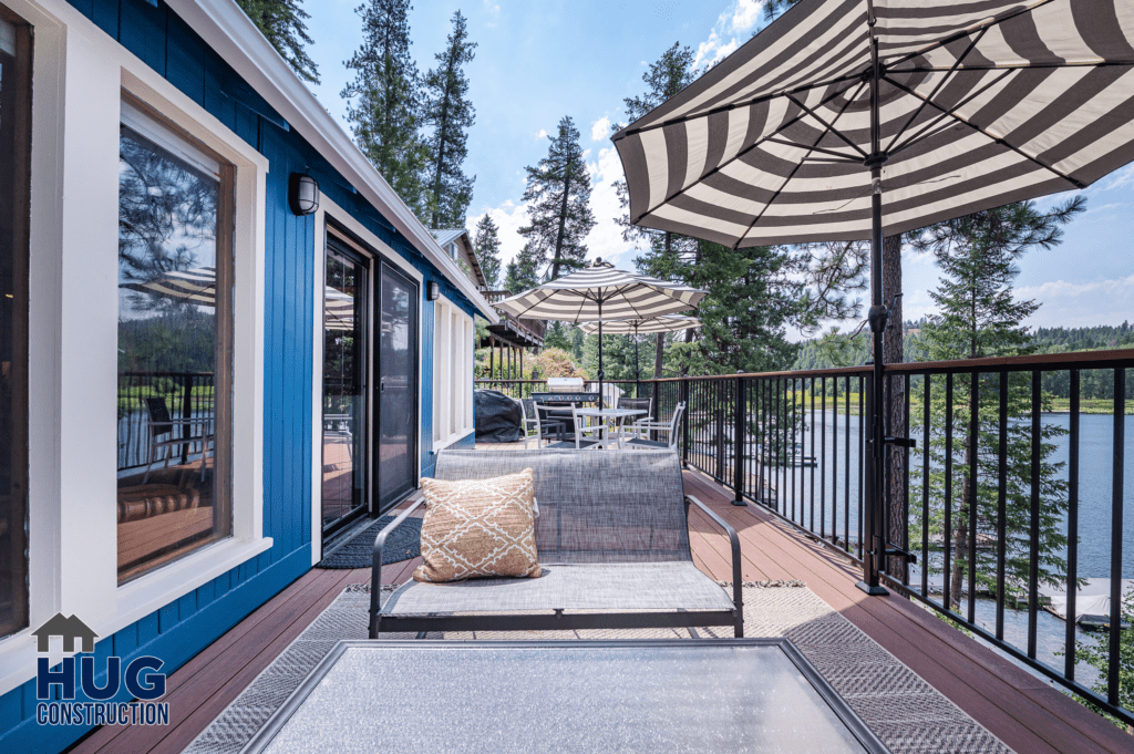 A spacious Silver Beach Cabin Remodel lakeside deck featuring modern outdoor furniture under striped umbrellas, with a tranquil water view and lush trees in the background.