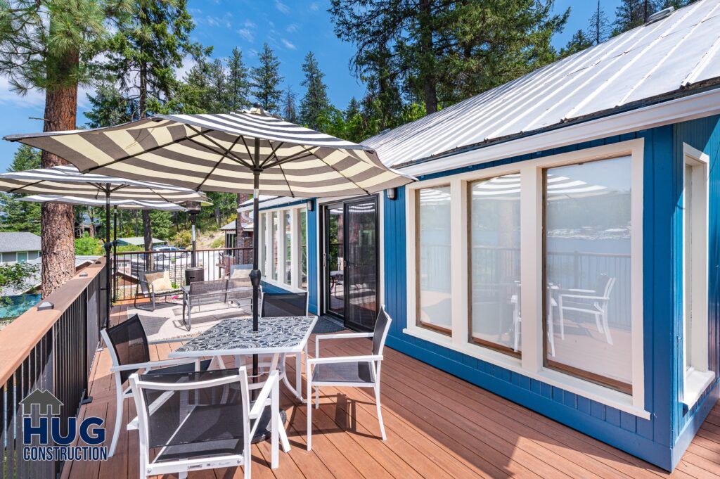 A sunny deck of a blue house with white trim features an open patio umbrella, outdoor furniture, and a view of trees and a lake, with a logo for "Silver Beach Cabin Remodel" in