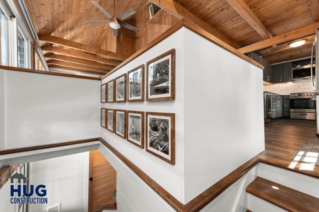 A modern Silver Beach Cabin Remodel kitchen interior featuring wooden beams on the ceiling, stainless steel appliances, a staircase to the left, and a series of framed pictures on the wall.