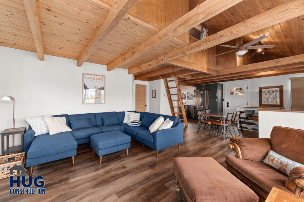 Silver Beach Cabin Remodel: Modern rustic living space with a blue sectional sofa, wooden beam ceilings, and an open-plan kitchen in the background.