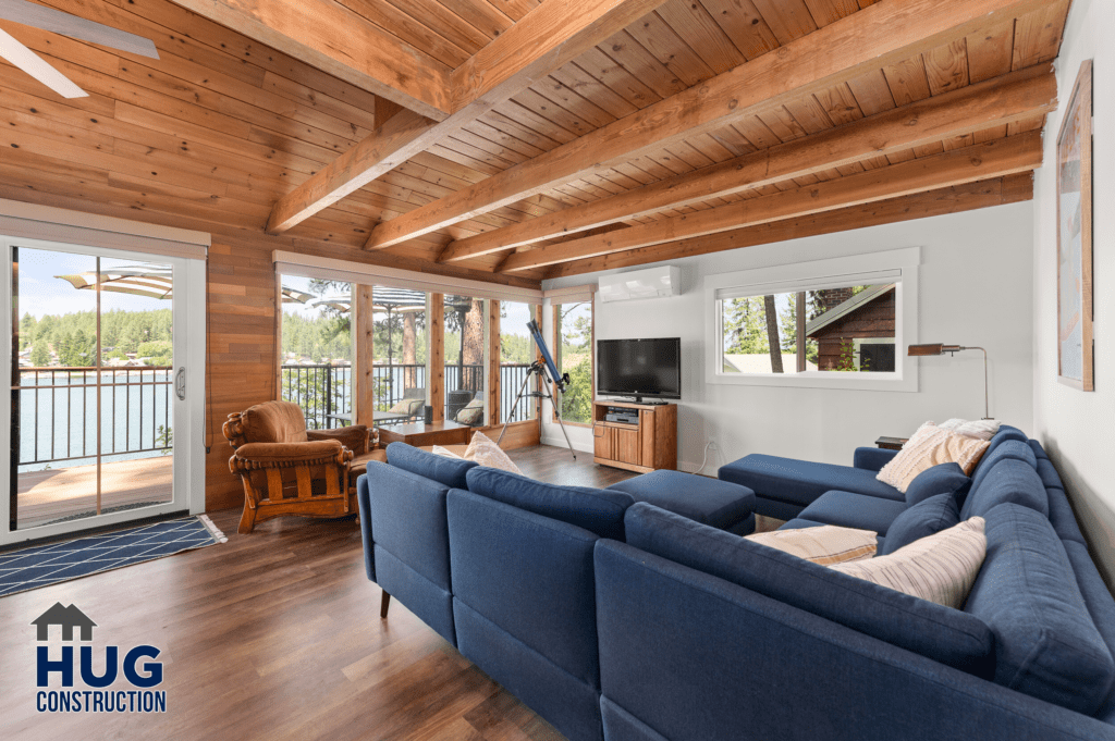 A modern Silver Beach Cabin living room featuring a blue sofa, wooden ceiling beams, hardwood floors, and a view of a lake through the balcony doors.