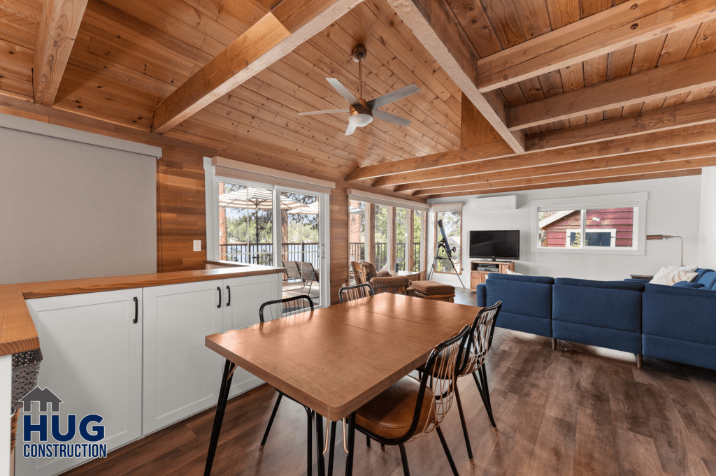Open-plan living space featuring a wooden dining table, white kitchen cabinetry, exposed beams, and a view through large glass doors to an outdoor area of the Silver Beach Cabin Remodel.
