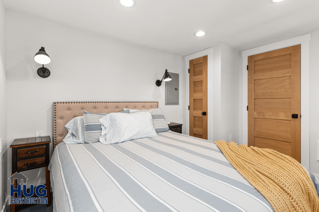 A neatly arranged bedroom in the Silver Beach Cabin Remodel, featuring a striped bedspread, a tufted headboard, two wooden doors, and wall-mounted reading lamps.