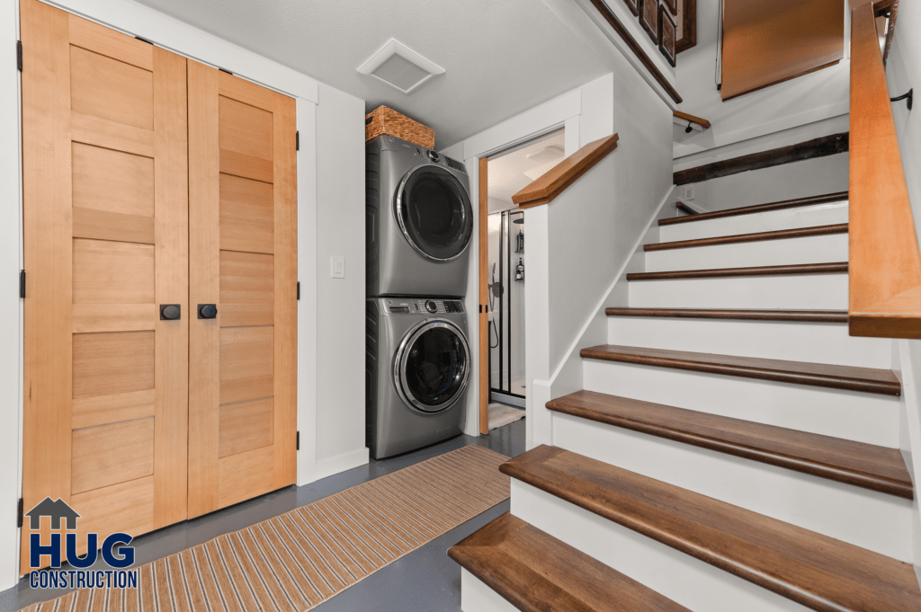 A modern laundry room from the Silver Beach Cabin Remodel with a stacked washer and dryer, located under the stairs in a well-lit space with wooden doors and stair balustrade.