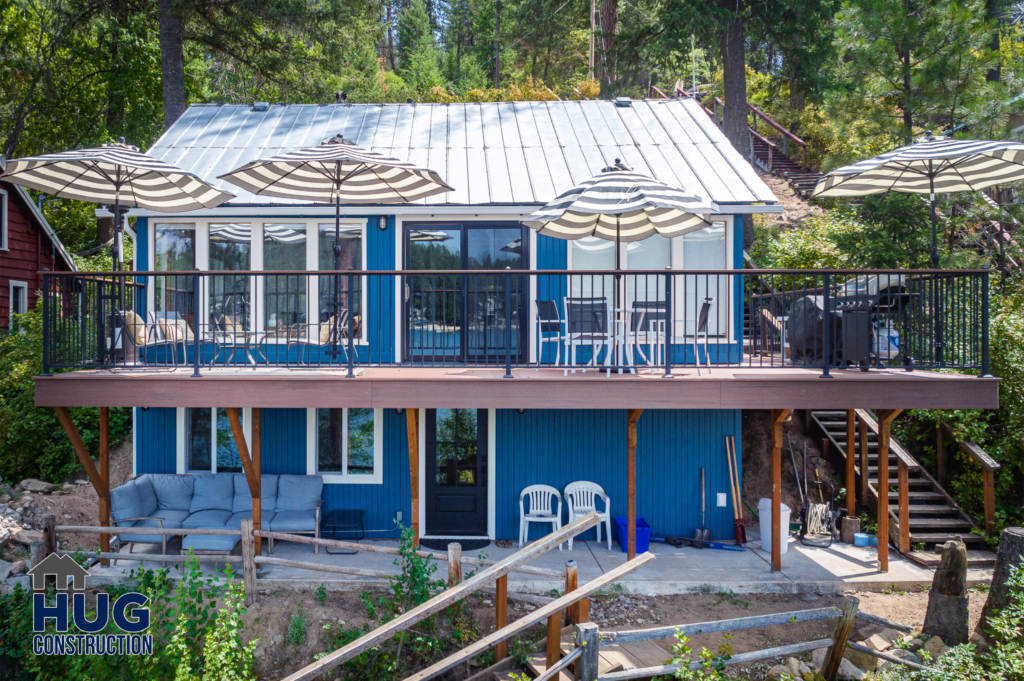 A Silver Beach Cabin Remodel: A two-story house with a blue exterior and a metal roof featuring a spacious upper deck with patio umbrellas and outdoor furniture.