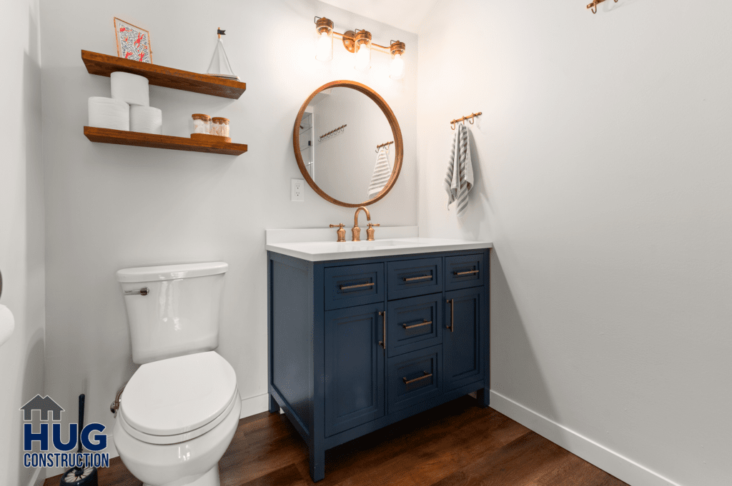 A modern bathroom in the Silver Beach Cabin Remodel with white walls featuring a blue vanity cabinet, a circular mirror, and floating wooden shelves.