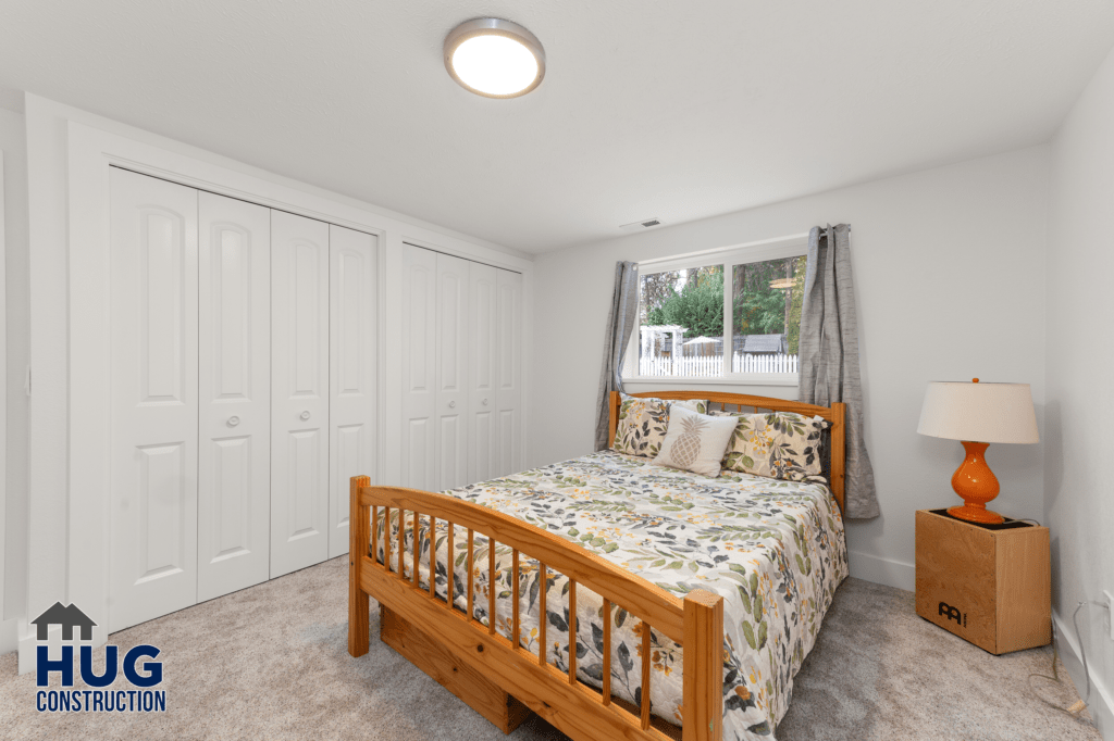 A neatly arranged bedroom featuring a wooden bed with patterned bedding, a large window with gray curtains, white closet doors, and a small bedside table with a lamp. A logo of 'Hug Construction