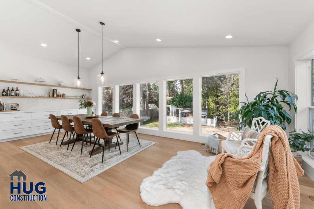 A spacious and modern kitchen dining area with white walls, a large window allowing natural light, and contemporary furniture, featuring a wooden table with surrounding chairs and a kitchen counter in the background after an extensive 