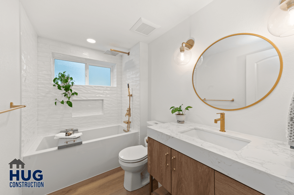 Modern bathroom interior, part of a 2-story addition, with white tiled walls, a wooden vanity with a white countertop, round mirror, brass fixtures, and a small window letting in natural light