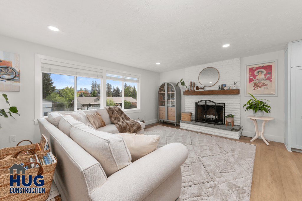 A bright, spacious living room with white walls, featuring a large sectional sofa, hardwood floors, a white brick fireplace, and several windows providing ample natural light as part of a 2-story addition.