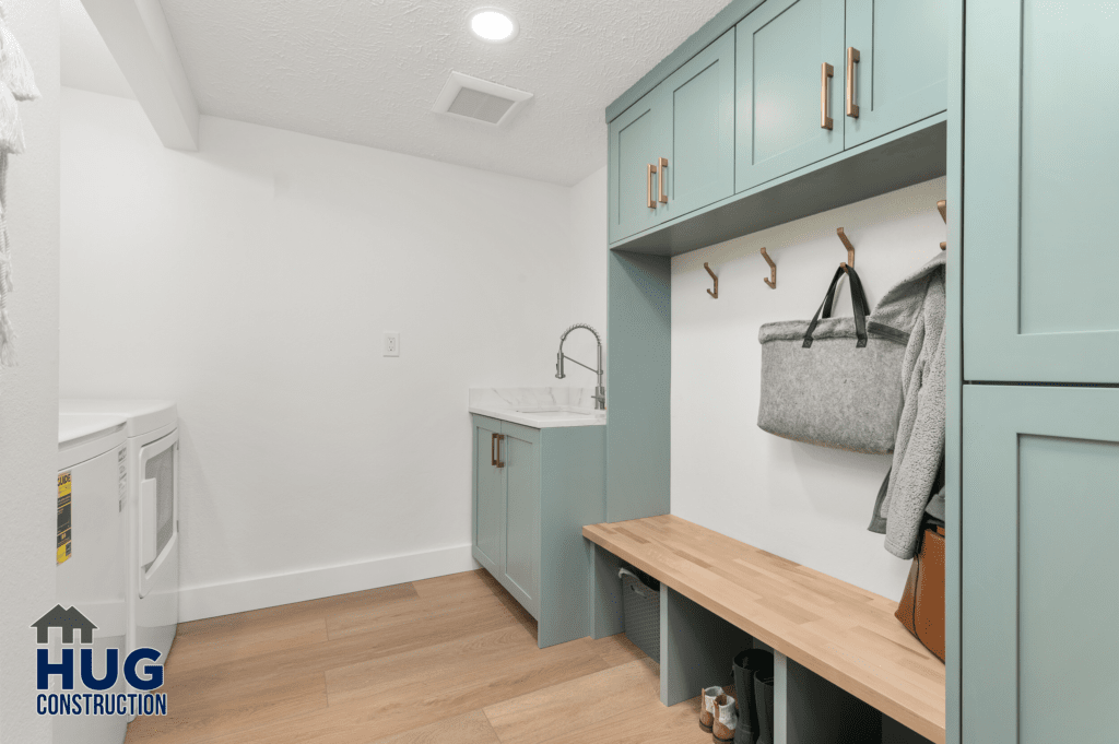 A modern 2-story addition laundry room featuring white walls, wooden flooring, teal cabinets, a sink, a washing machine, and a bench with coat hooks above it.