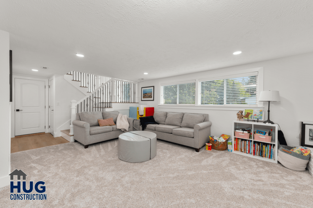 A neatly organized living room with two grey sofas, a white ottoman, a bookshelf, and a staircase leading to an upper level as part of a 2-story addition. The room is well
